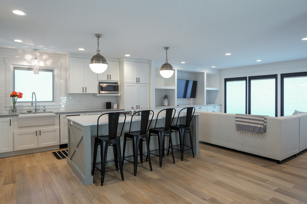 Design ideas for a medium sized nautical l-shaped open plan kitchen in Grand Rapids with a belfast sink, shaker cabinets, white cabinets, engineered stone countertops, white splashback, metro tiled splashback, stainless steel appliances, light hardwood flooring and an island.