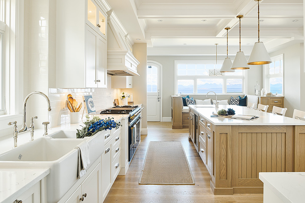 This is an example of a large nautical l-shaped kitchen/diner in Vancouver with a belfast sink, shaker cabinets, white cabinets, granite worktops, white splashback, ceramic splashback, coloured appliances, light hardwood flooring, an island, beige floors and white worktops.