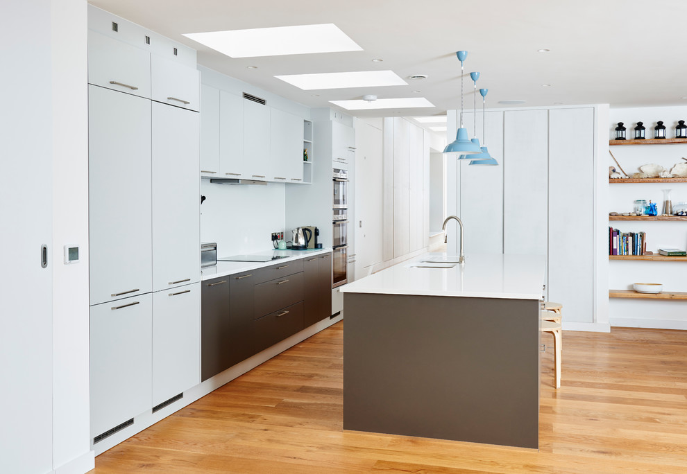 Medium sized contemporary galley open plan kitchen in Devon with an integrated sink, flat-panel cabinets, white cabinets, white splashback, stainless steel appliances, medium hardwood flooring and an island.