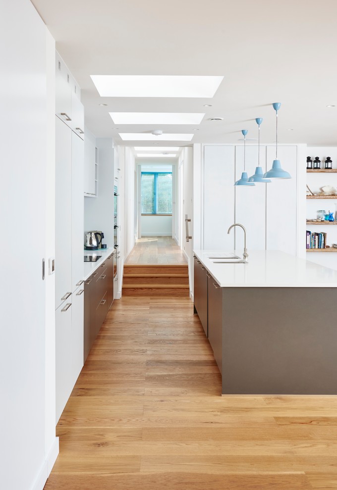 На фото: параллельная кухня-гостиная среднего размера в современном стиле с монолитной мойкой, плоскими фасадами, белыми фасадами, паркетным полом среднего тона и островом