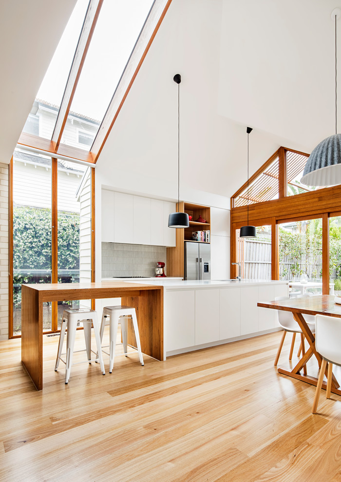 Zweizeilige Moderne Wohnküche mit flächenbündigen Schrankfronten, weißen Schränken, Küchenrückwand in Grau, Küchengeräten aus Edelstahl, hellem Holzboden und Kücheninsel in Melbourne
