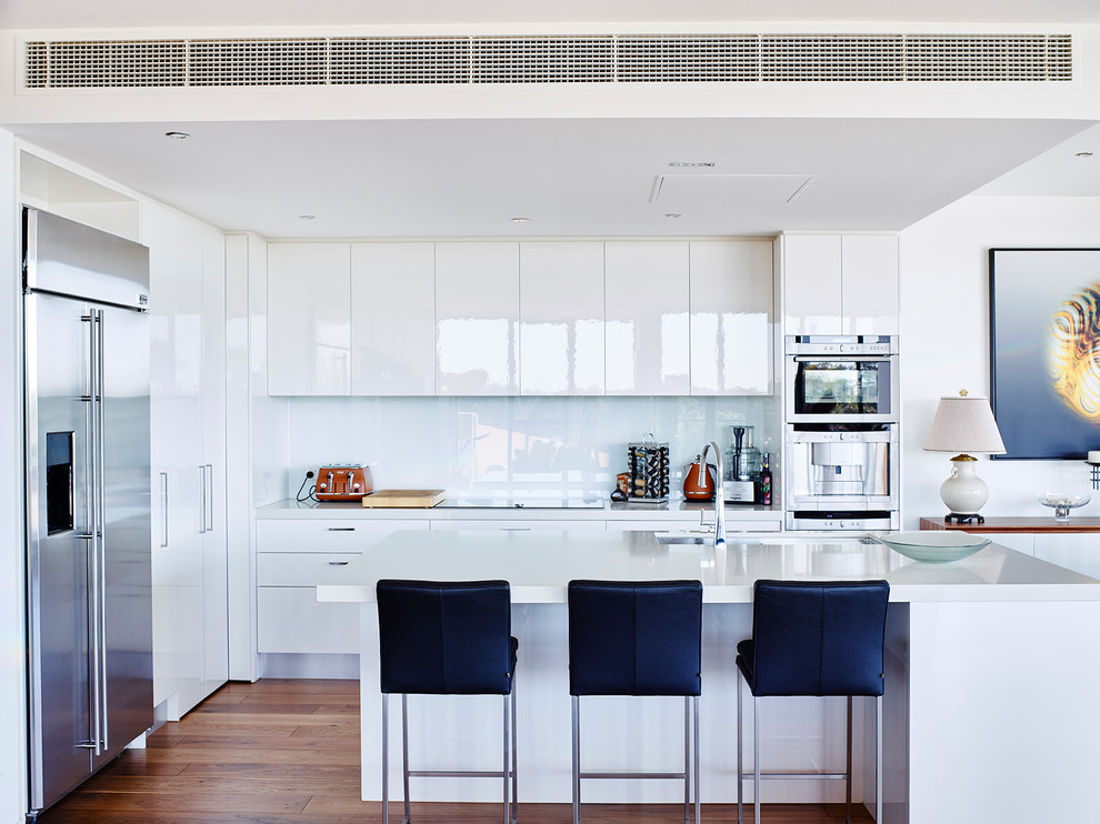 Modern inredning av ett kök, med en undermonterad diskho, släta luckor, vita skåp, vitt stänkskydd, glaspanel som stänkskydd, rostfria vitvaror, mellanmörkt trägolv och en köksö