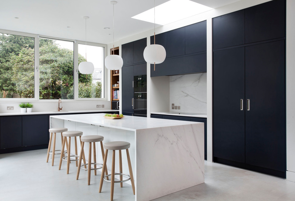 Modern inredning av ett vit vitt kök, med släta luckor, blå skåp, marmorbänkskiva, vitt stänkskydd, stänkskydd i marmor, svarta vitvaror, betonggolv, en köksö och grått golv