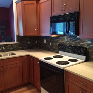Bild på ett avskilt, litet funkis l-kök, med en undermonterad diskho, skåp i shakerstil, bruna skåp, bänkskiva i kvartsit, grönt stänkskydd, stänkskydd i stenkakel, svarta vitvaror, ljust trägolv och en köksö