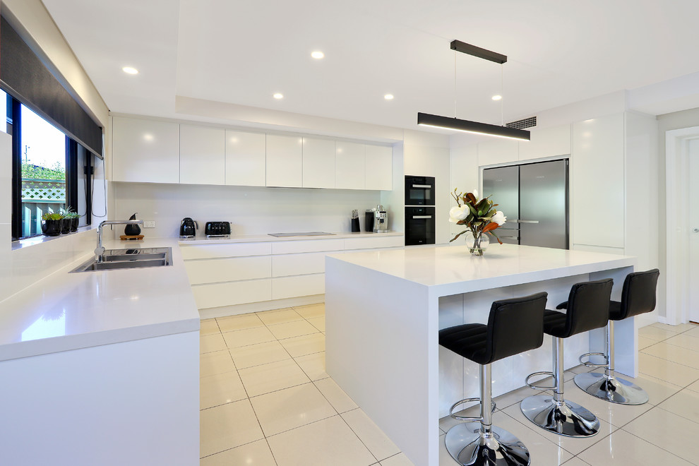 Example of a large minimalist u-shaped porcelain tile and beige floor enclosed kitchen design in Sydney with a drop-in sink, flat-panel cabinets, white cabinets, quartz countertops, white backsplash, stone slab backsplash, black appliances, an island and white countertops