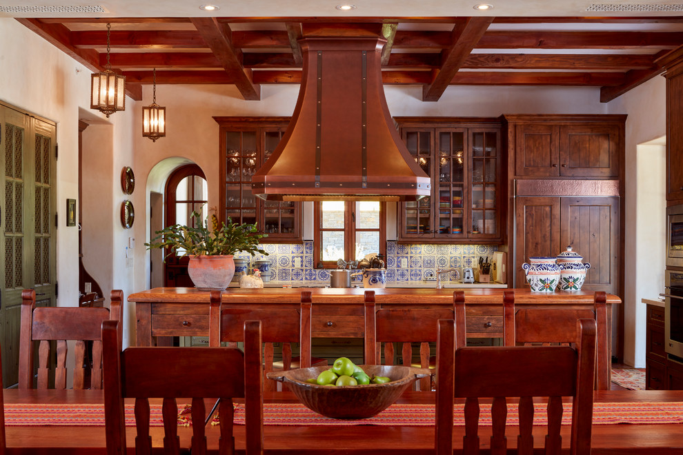Пример оригинального дизайна: прямая кухня в средиземноморском стиле с обеденным столом, фасадами с утопленной филенкой, фасадами цвета дерева среднего тона, деревянной столешницей, синим фартуком, техникой под мебельный фасад и островом