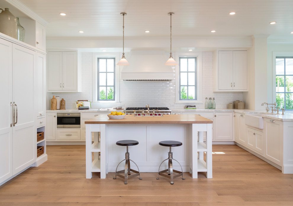 Photo of a classic kitchen in Los Angeles with a belfast sink, shaker cabinets, white cabinets, white splashback, metro tiled splashback, stainless steel appliances, light hardwood flooring and an island.