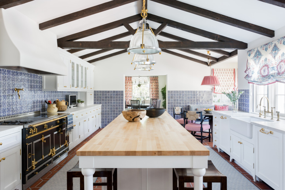 Example of a mid-sized tuscan u-shaped terra-cotta tile, red floor and vaulted ceiling eat-in kitchen design in Los Angeles with a farmhouse sink, flat-panel cabinets, white cabinets, marble countertops, blue backsplash, ceramic backsplash, black appliances, an island and white countertops