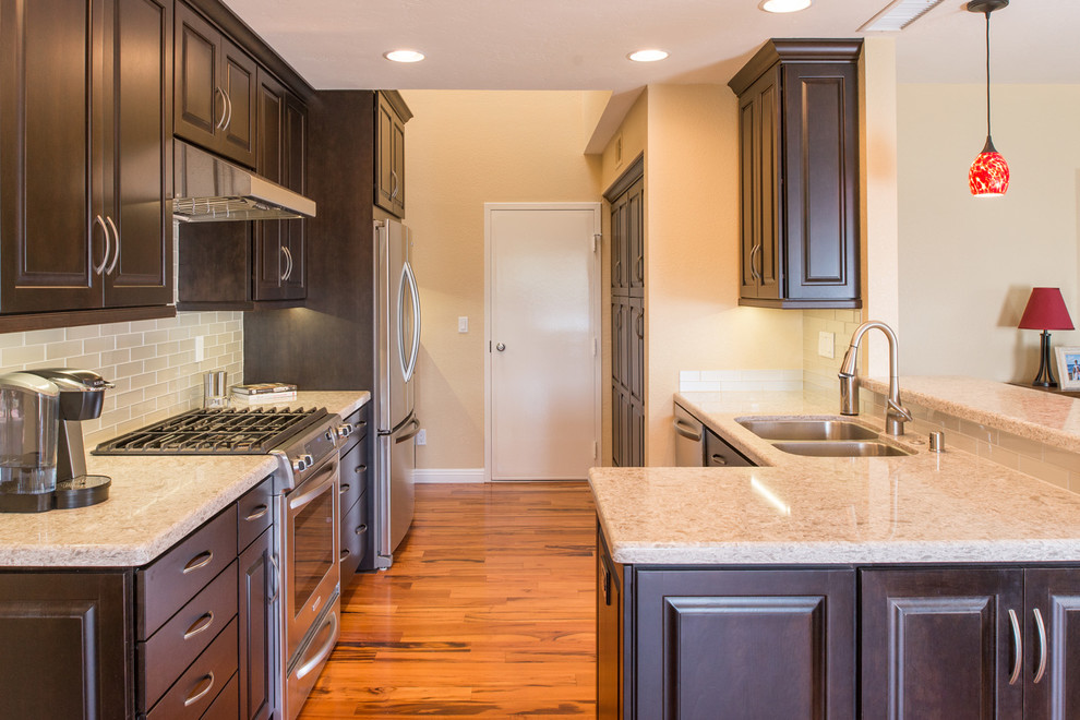 Small elegant u-shaped medium tone wood floor eat-in kitchen photo in San Diego with a drop-in sink, raised-panel cabinets, dark wood cabinets, granite countertops, white backsplash, glass tile backsplash, stainless steel appliances and a peninsula