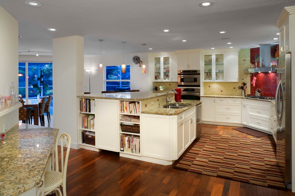 Inspiration for a mid-sized eclectic l-shaped medium tone wood floor open concept kitchen remodel in Tampa with a double-bowl sink, shaker cabinets, white cabinets, granite countertops, beige backsplash, glass tile backsplash, stainless steel appliances and an island