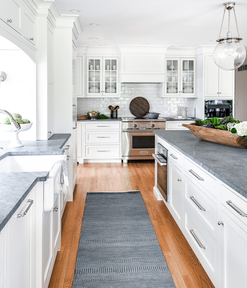 Kitchen - large transitional medium tone wood floor and brown floor kitchen idea in Philadelphia with a farmhouse sink, white cabinets, soapstone countertops, white backsplash, ceramic backsplash, paneled appliances, an island, gray countertops and shaker cabinets