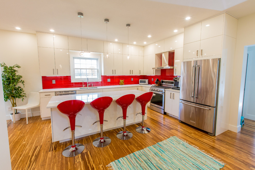 Bild på ett stort funkis vit vitt kök, med en undermonterad diskho, släta luckor, vita skåp, bänkskiva i kvartsit, rött stänkskydd, stänkskydd i glaskakel, rostfria vitvaror, bambugolv, en köksö och brunt golv