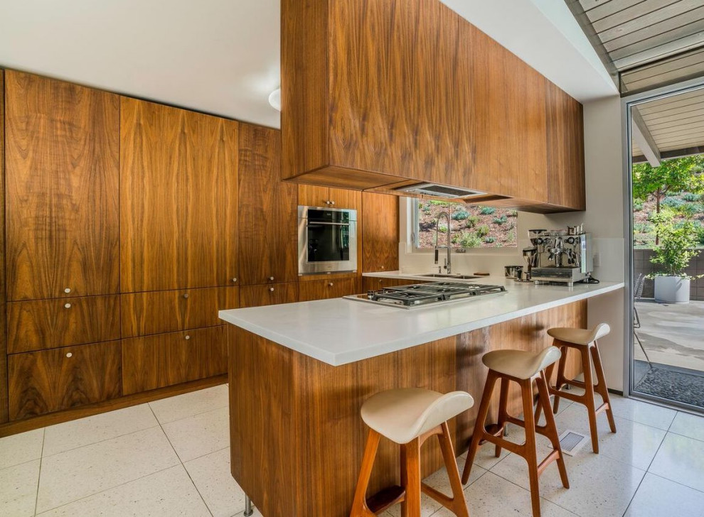 Example of a mid-sized 1950s terrazzo floor, white floor and exposed beam kitchen design in Los Angeles with a double-bowl sink, flat-panel cabinets, solid surface countertops, stainless steel appliances, a peninsula, white countertops and medium tone wood cabinets