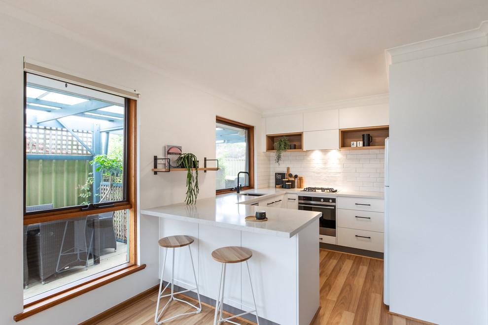 Inspiration for a scandinavian u-shaped kitchen in Adelaide with a submerged sink, flat-panel cabinets, white cabinets, engineered stone countertops, white splashback, metro tiled splashback, white appliances, brown floors, white worktops and laminate floors.