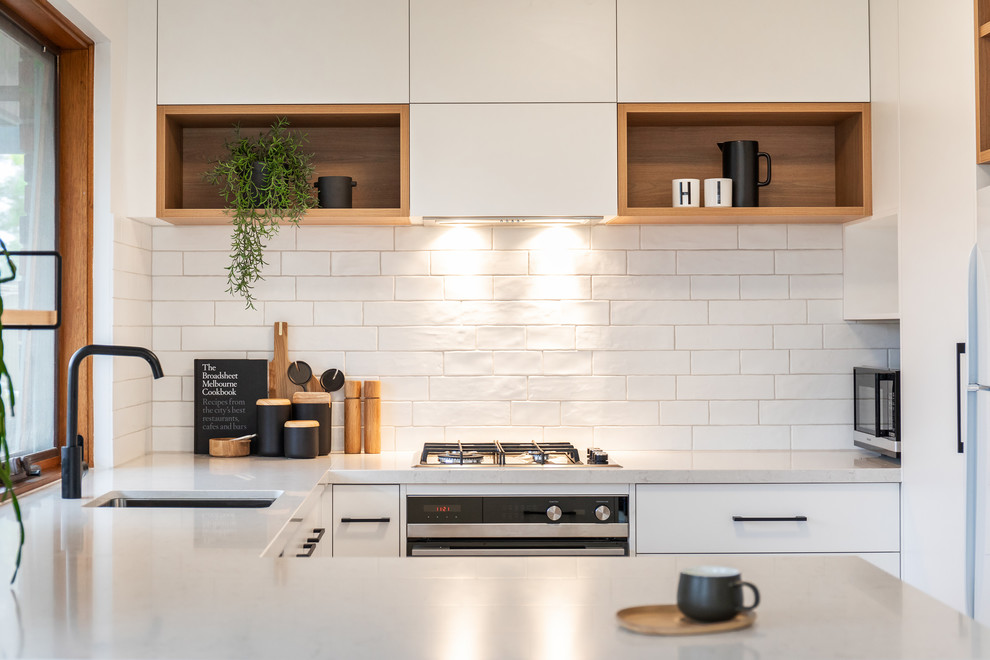 This is an example of a scandinavian u-shaped kitchen in Adelaide with a submerged sink, flat-panel cabinets, white cabinets, engineered stone countertops, white splashback, metro tiled splashback, white appliances, laminate floors, brown floors and white worktops.