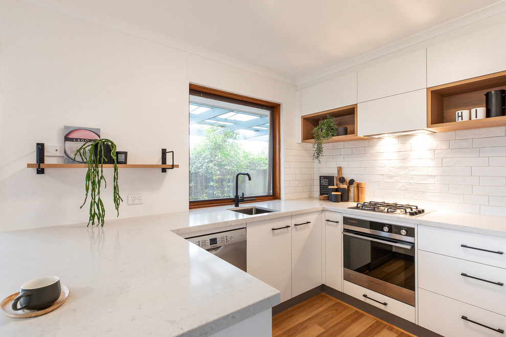 Example of a danish u-shaped brown floor and laminate floor kitchen design in Adelaide with an undermount sink, flat-panel cabinets, white cabinets, quartz countertops, white backsplash, subway tile backsplash, white appliances and white countertops