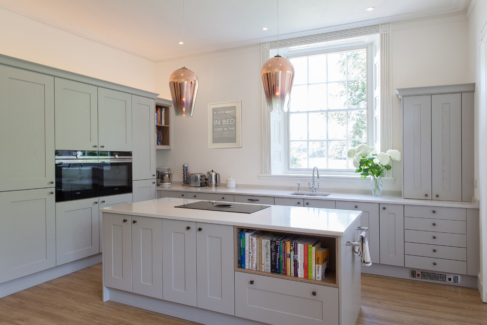 Medium sized traditional l-shaped kitchen in Hampshire with a built-in sink, grey cabinets, black appliances, an island, recessed-panel cabinets, quartz worktops, light hardwood flooring and brown floors.