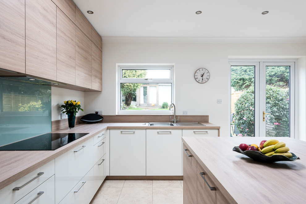 Design ideas for a scandinavian l-shaped kitchen/diner in Surrey with a single-bowl sink, flat-panel cabinets, light wood cabinets, laminate countertops, green splashback, glass sheet splashback, black appliances, ceramic flooring, an island, beige floors and beige worktops.