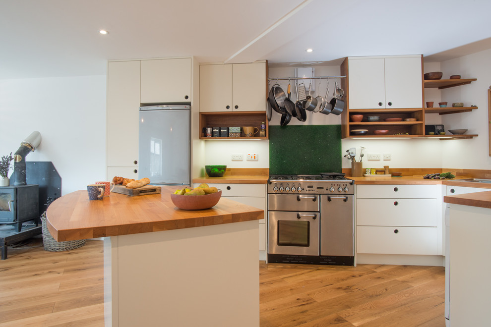 Foto på ett mellanstort minimalistiskt kök, med en dubbel diskho, släta luckor, vita skåp, träbänkskiva, grönt stänkskydd, glaspanel som stänkskydd, rostfria vitvaror, ljust trägolv och en köksö
