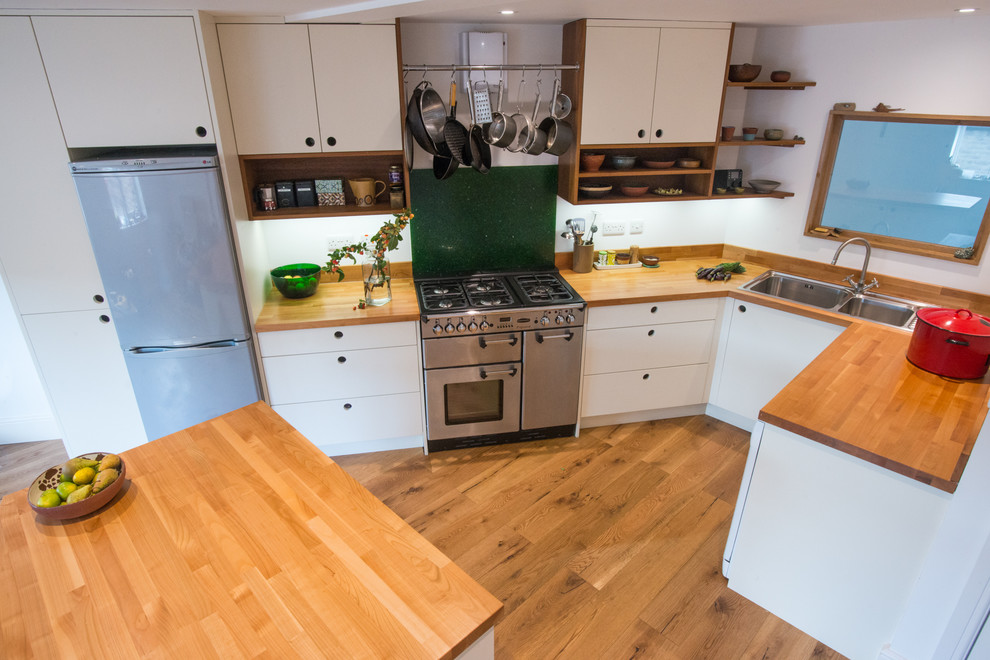 Design ideas for a medium sized scandinavian u-shaped open plan kitchen in Other with a double-bowl sink, flat-panel cabinets, white cabinets, wood worktops, green splashback, glass sheet splashback, stainless steel appliances, light hardwood flooring and an island.