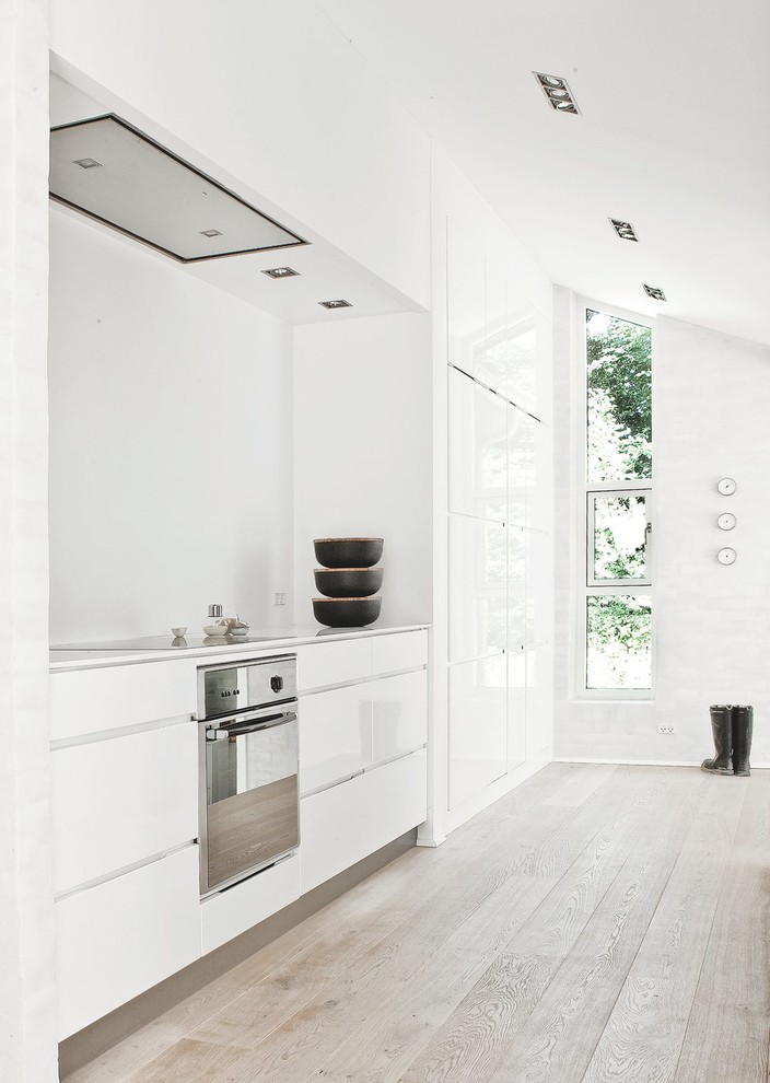 Imagen de cocina lineal nórdica de tamaño medio cerrada sin isla con armarios con paneles lisos, puertas de armario blancas, salpicadero blanco, electrodomésticos de acero inoxidable, suelo de madera clara y encimera de laminado