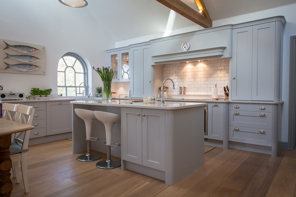 Inspiration for a mid-sized cottage l-shaped medium tone wood floor and brown floor eat-in kitchen remodel in Hertfordshire with a farmhouse sink, shaker cabinets, gray cabinets, quartz countertops, white backsplash, subway tile backsplash, stainless steel appliances, an island and gray countertops
