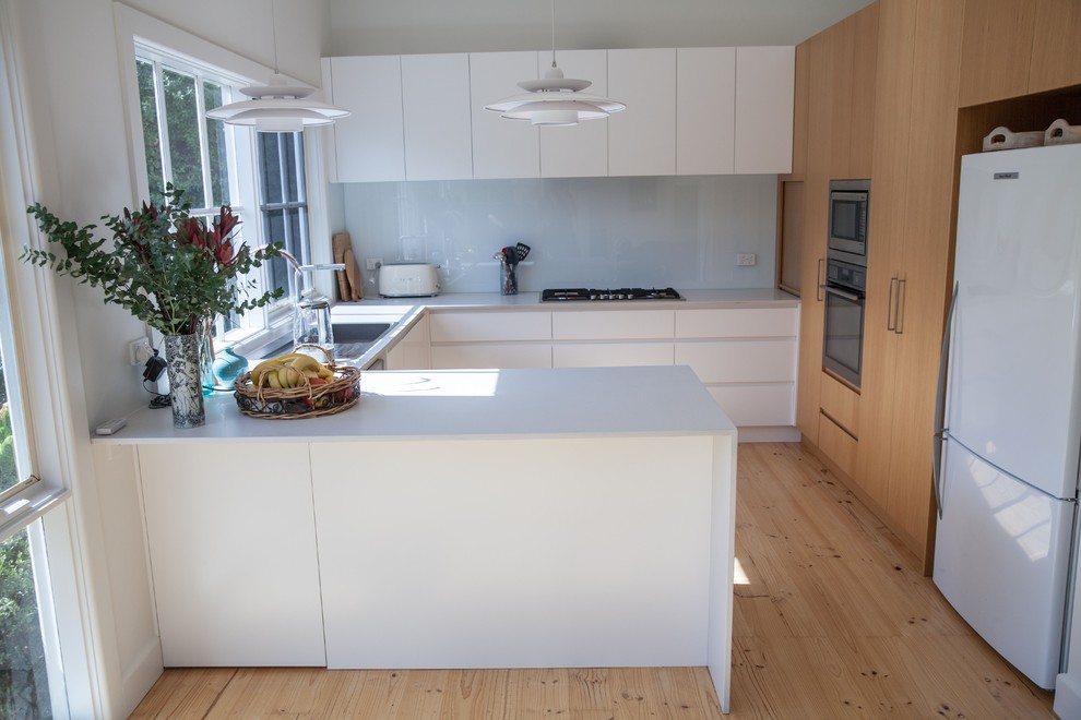 Inredning av ett minimalistiskt mellanstort grå grått kök, med en undermonterad diskho, släta luckor, vita skåp, granitbänkskiva, vitt stänkskydd, glaspanel som stänkskydd, ljust trägolv, en halv köksö och rostfria vitvaror