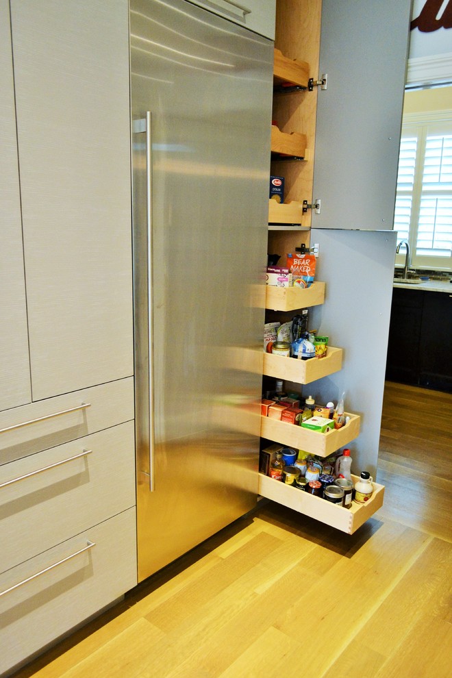 This is an example of a large contemporary l-shaped kitchen pantry in New York with a submerged sink, glass-front cabinets, light wood cabinets, white splashback, glass tiled splashback, stainless steel appliances, light hardwood flooring and an island.