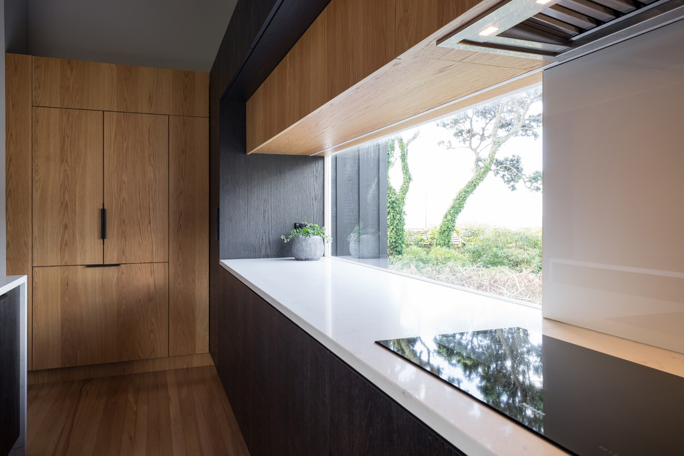 Mittelgroße Moderne Wohnküche in L-Form mit Quarzwerkstein-Arbeitsplatte, Küchenrückwand in Weiß, Glasrückwand, Elektrogeräten mit Frontblende, braunem Holzboden, Kücheninsel, Unterbauwaschbecken, flächenbündigen Schrankfronten, hellbraunen Holzschränken und braunem Boden in Auckland