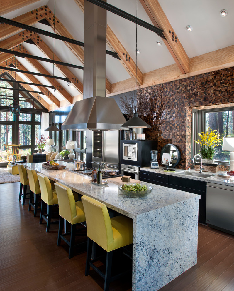 Bild på ett stort industriellt kök, med en undermonterad diskho, släta luckor, svarta skåp, brunt stänkskydd, rostfria vitvaror, mellanmörkt trägolv, en köksö och stänkskydd i mosaik