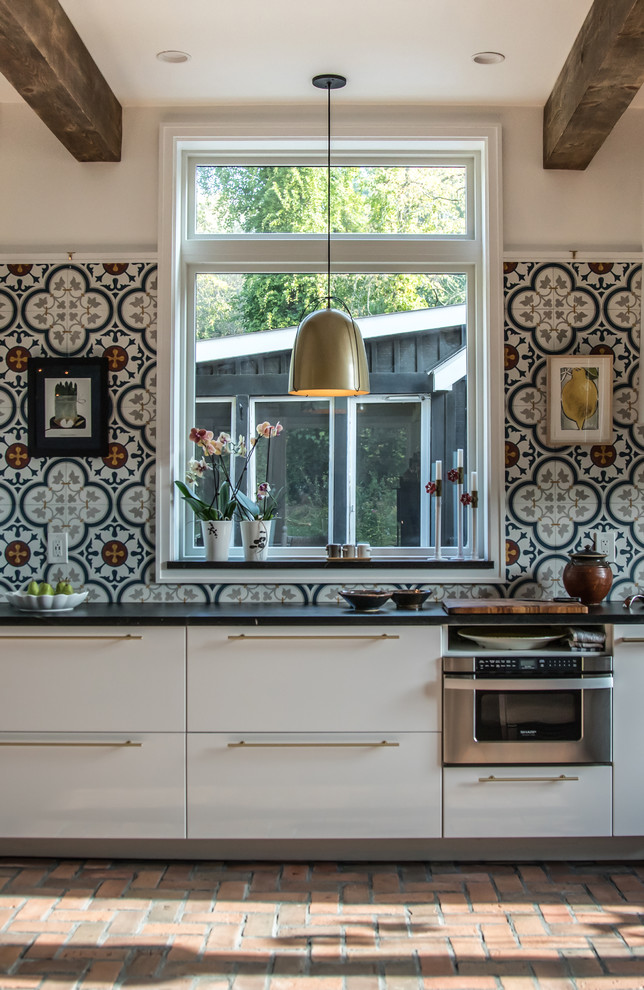 Bild på ett mellanstort lantligt kök, med en undermonterad diskho, släta luckor, vita skåp, bänkskiva i täljsten, flerfärgad stänkskydd, stänkskydd i cementkakel, rostfria vitvaror, tegelgolv och rött golv
