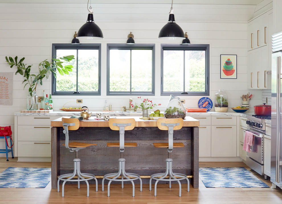 Réalisation d'une cuisine champêtre en L avec un placard à porte shaker, des portes de placard blanches, un électroménager en acier inoxydable, parquet clair, îlot et fenêtre au-dessus de l'évier.