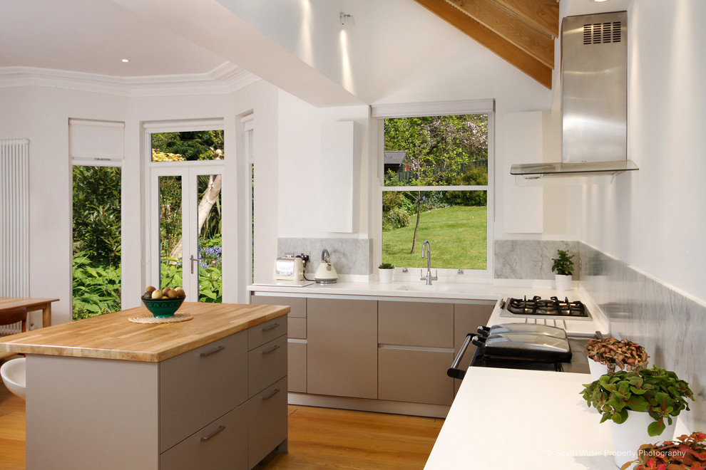 This is an example of a medium sized contemporary l-shaped open plan kitchen in Other with an island, an integrated sink, flat-panel cabinets, beige cabinets, composite countertops, grey splashback, marble splashback, black appliances, medium hardwood flooring, brown floors, white worktops and exposed beams.
