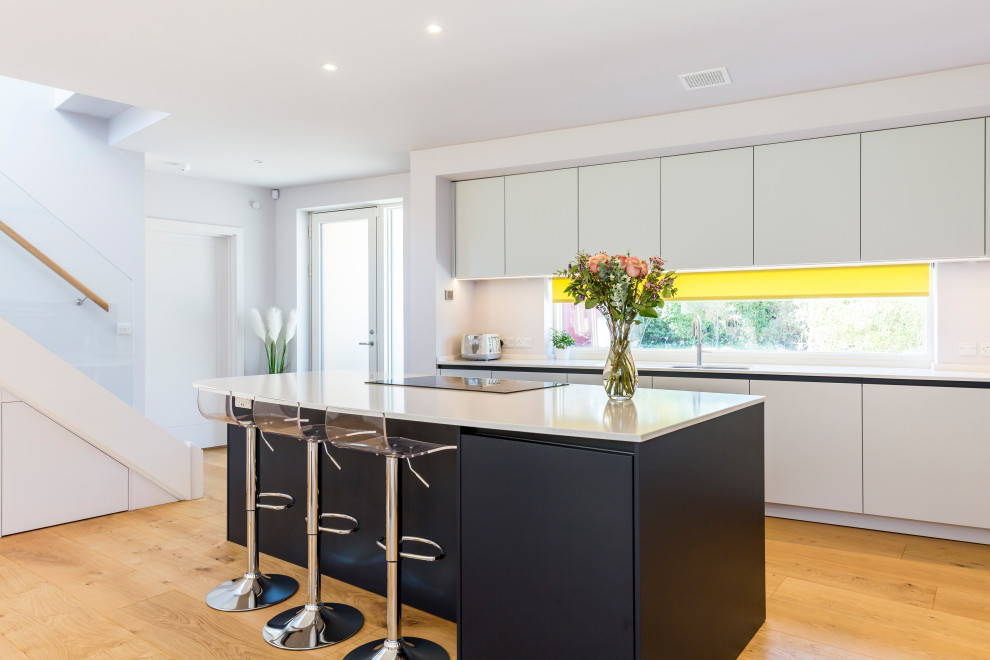Moderne Küche mit Unterbauwaschbecken, flächenbündigen Schrankfronten, weißen Schränken, Rückwand-Fenster, hellem Holzboden, Kücheninsel und weißer Arbeitsplatte in Sussex