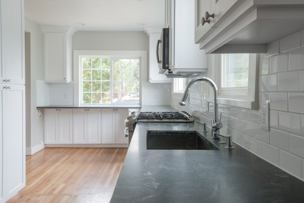 Geschlossene, Mittelgroße Klassische Küche in L-Form mit Unterbauwaschbecken, Schrankfronten im Shaker-Stil, weißen Schränken, Speckstein-Arbeitsplatte, Küchenrückwand in Weiß, Rückwand aus Metrofliesen, Küchengeräten aus Edelstahl, hellem Holzboden und Kücheninsel in Sonstige