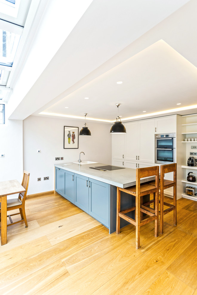 Medium sized classic galley open plan kitchen in London with a belfast sink, shaker cabinets, stainless steel appliances, light hardwood flooring and a breakfast bar.