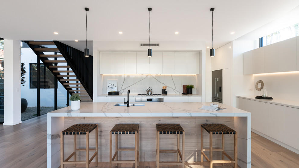 Inspiration for a contemporary l-shaped kitchen in Other with white cabinets, marble worktops, marble splashback, stainless steel appliances, laminate floors, an island, a submerged sink, flat-panel cabinets, white splashback, brown floors and white worktops.