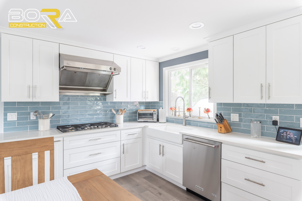This is an example of a medium sized traditional l-shaped kitchen/diner in Seattle with a belfast sink, shaker cabinets, white cabinets, engineered stone countertops, blue splashback, ceramic splashback, stainless steel appliances, light hardwood flooring, no island, grey floors and white worktops.