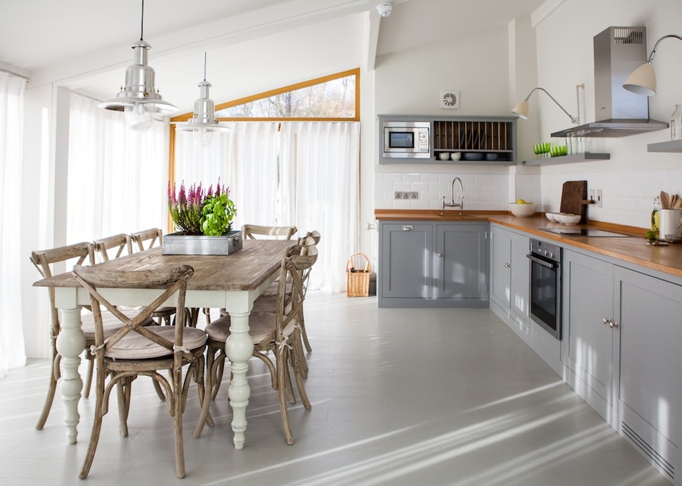 Réalisation d'une cuisine américaine champêtre avec un placard à porte shaker, des portes de placard grises, un plan de travail en bois, une crédence blanche, une crédence en céramique, un électroménager en acier inoxydable, un évier de ferme, parquet peint et aucun îlot.