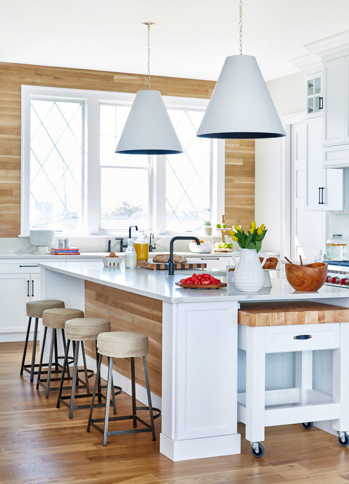 Example of a beach style light wood floor and beige floor kitchen design in New York with shaker cabinets, white cabinets, wood backsplash, stainless steel appliances, white countertops, beige backsplash, marble countertops and an island