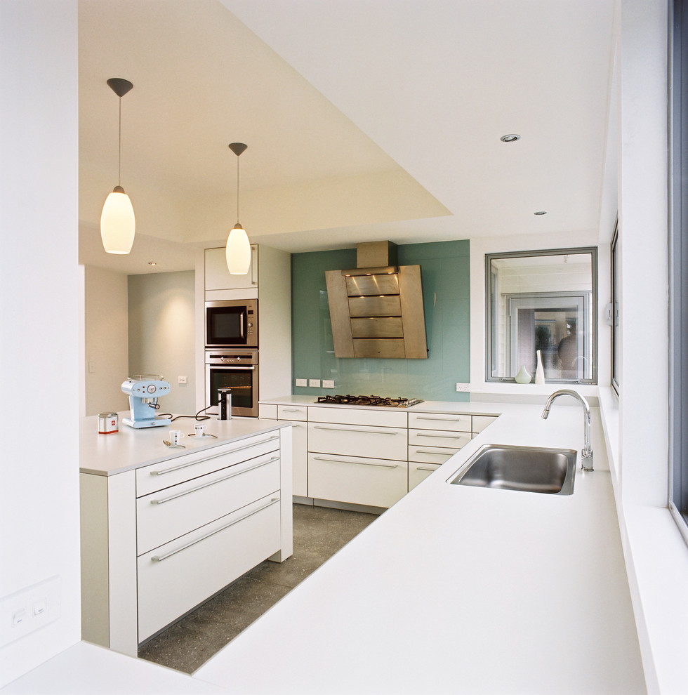 ウェリントンにあるコンテンポラリースタイルのおしゃれなキッチン (シングルシンク、白いキャビネット、ラミネートカウンター、青いキッチンパネル、ガラス板のキッチンパネル、シルバーの調理設備、コンクリートの床) の写真