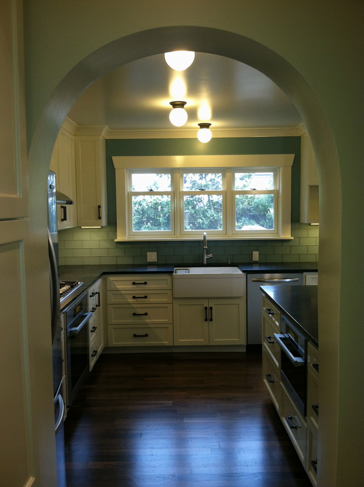 Example of a large farmhouse l-shaped dark wood floor open concept kitchen design in Seattle with a farmhouse sink, shaker cabinets, white cabinets, soapstone countertops, green backsplash, subway tile backsplash, stainless steel appliances and an island