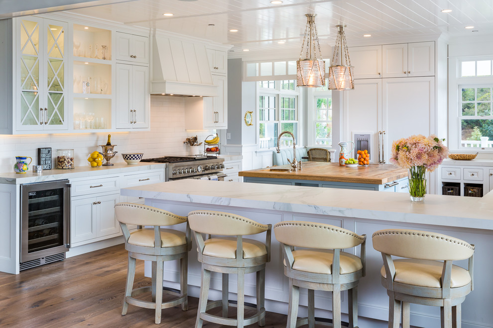 This is an example of a large beach style kitchen/diner in San Diego with a belfast sink, beaded cabinets, white cabinets, composite countertops, white splashback, ceramic splashback, integrated appliances, medium hardwood flooring, an island and grey floors.