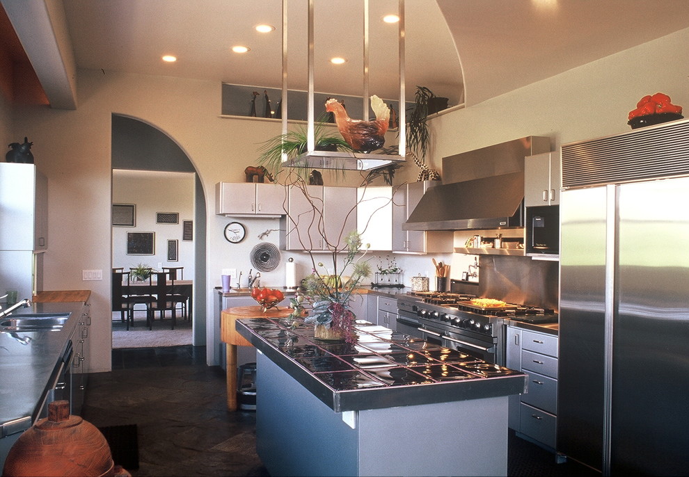 Large industrial l-shaped enclosed kitchen in Phoenix with flat-panel cabinets, grey cabinets, tile countertops, an island, a double-bowl sink, grey splashback, stainless steel appliances, slate flooring, brown floors and black worktops.