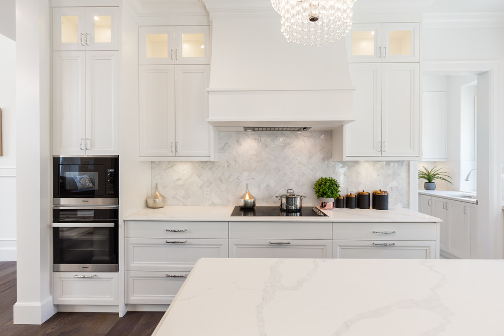 Inredning av ett klassiskt mellanstort vit vitt kök, med en undermonterad diskho, luckor med infälld panel, vita skåp, bänkskiva i kvarts, grått stänkskydd, stänkskydd i marmor, integrerade vitvaror, mellanmörkt trägolv, en köksö och brunt golv