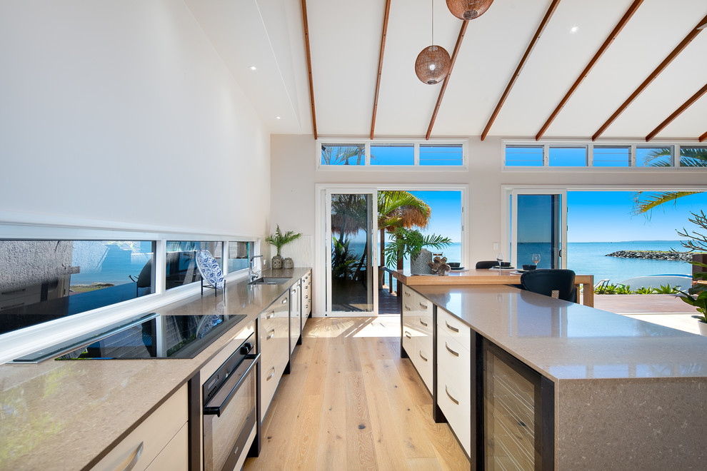 Exemple d'une cuisine parallèle bord de mer avec un évier encastré, un placard à porte plane, des portes de placard blanches, parquet clair, îlot, un sol beige et fenêtre.