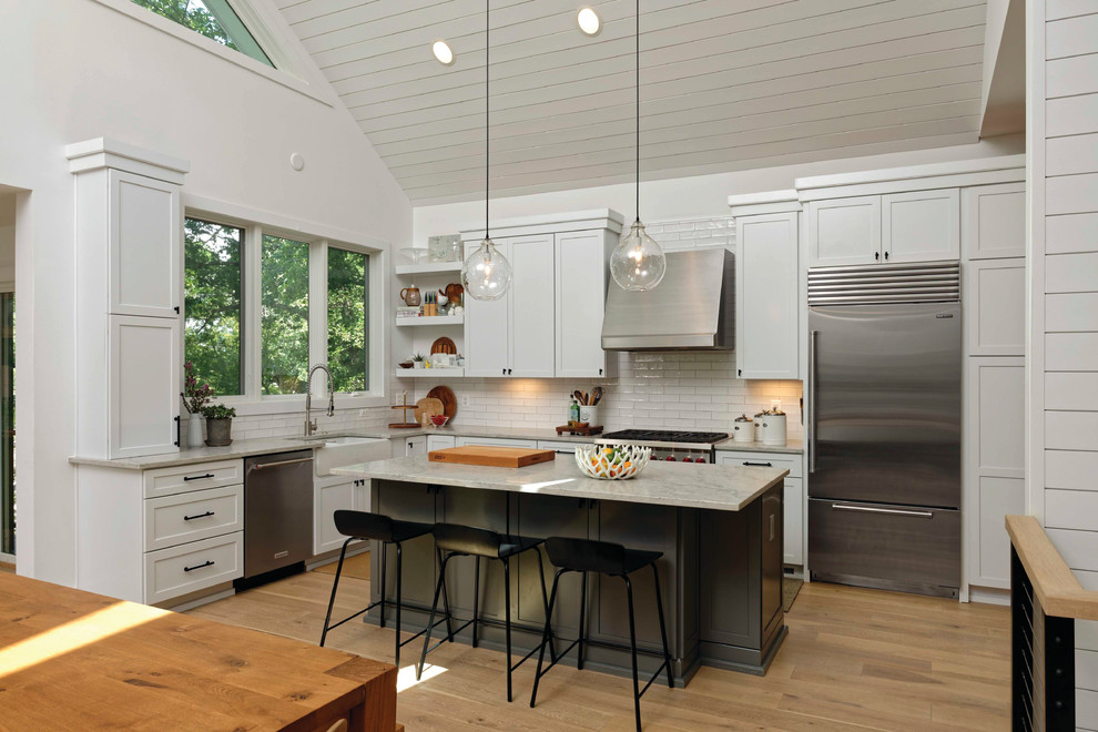 This is an example of a country l-shaped kitchen/diner in Baltimore with a belfast sink, shaker cabinets, white cabinets, white splashback, metro tiled splashback, stainless steel appliances, light hardwood flooring, an island, beige floors and grey worktops.