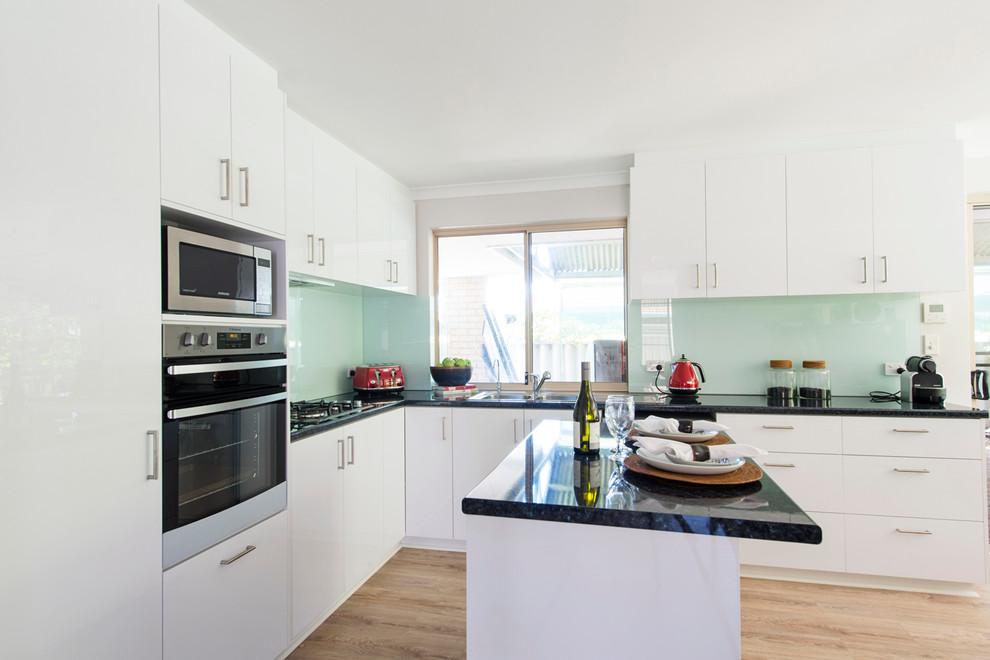 Mid-sized minimalist l-shaped light wood floor and brown floor eat-in kitchen photo in Perth with a double-bowl sink, flat-panel cabinets, white cabinets, laminate countertops, green backsplash, glass sheet backsplash, stainless steel appliances, an island and black countertops