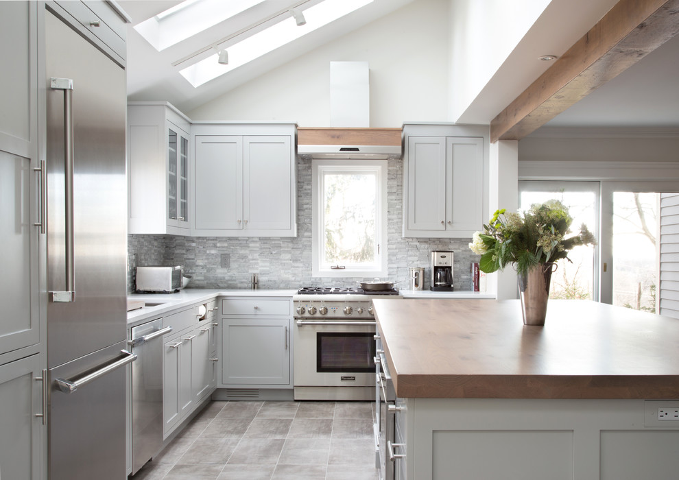 Inspiration for a mid-sized transitional l-shaped porcelain tile open concept kitchen remodel in New York with an undermount sink, shaker cabinets, gray cabinets, wood countertops, gray backsplash, stone tile backsplash, stainless steel appliances and an island