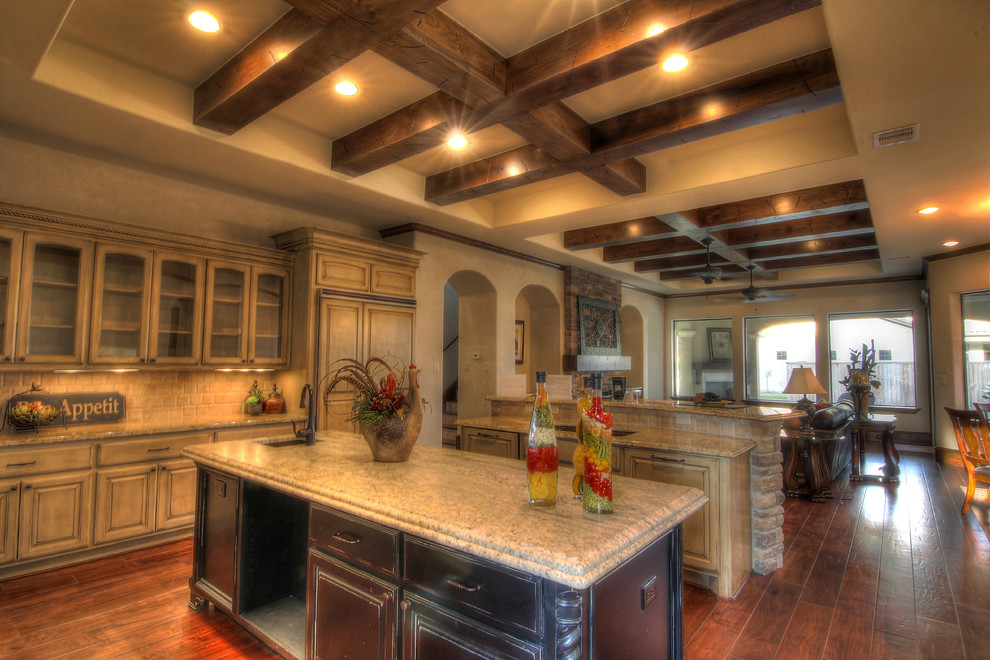 Example of a large tuscan l-shaped dark wood floor and brown floor open concept kitchen design in Houston with an undermount sink, raised-panel cabinets, distressed cabinets, granite countertops, beige backsplash, stone slab backsplash, paneled appliances and two islands
